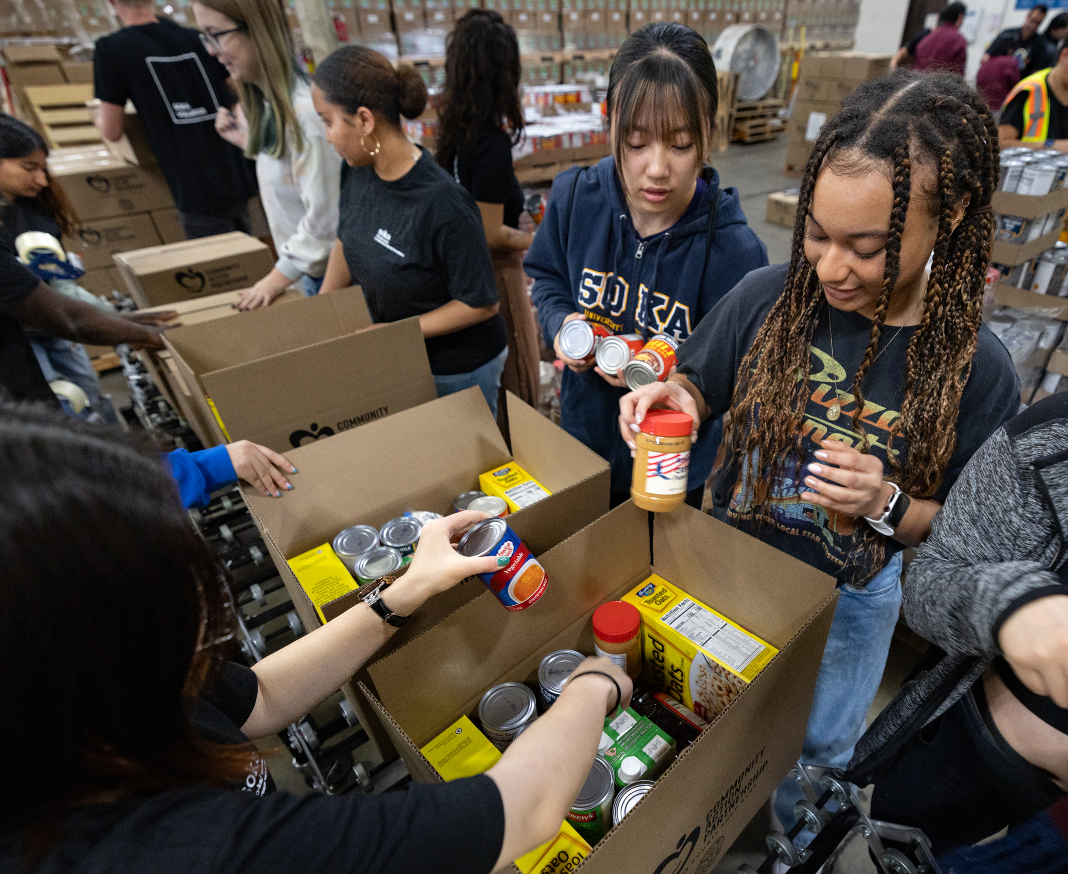 A group of young volunteers, including a student in a Soka University hoodie, are busy packing cardboard boxes with non-perishable food items like canned soup, peanut butter, and cereal at a large food bank warehouse.