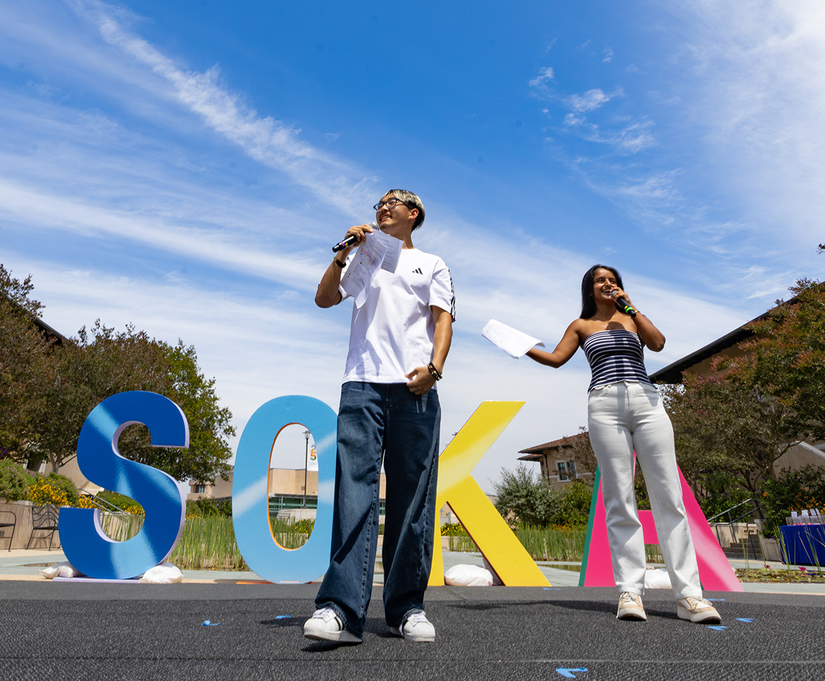 Two students stand outdoors on a stage in front of large, colorful letters spelling "SOKA." Both hold microphones and papers while addressing an audience under a bright blue sky with wispy clouds.