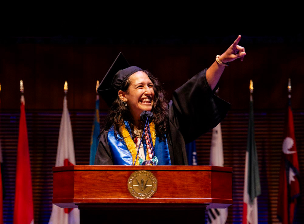 A smiling graduate stands behind a wooden podium adorned with a university seal and a large bouquet of colorful flowers. She wears a graduation cap and blue stole, happily pointing toward the audience with one hand.