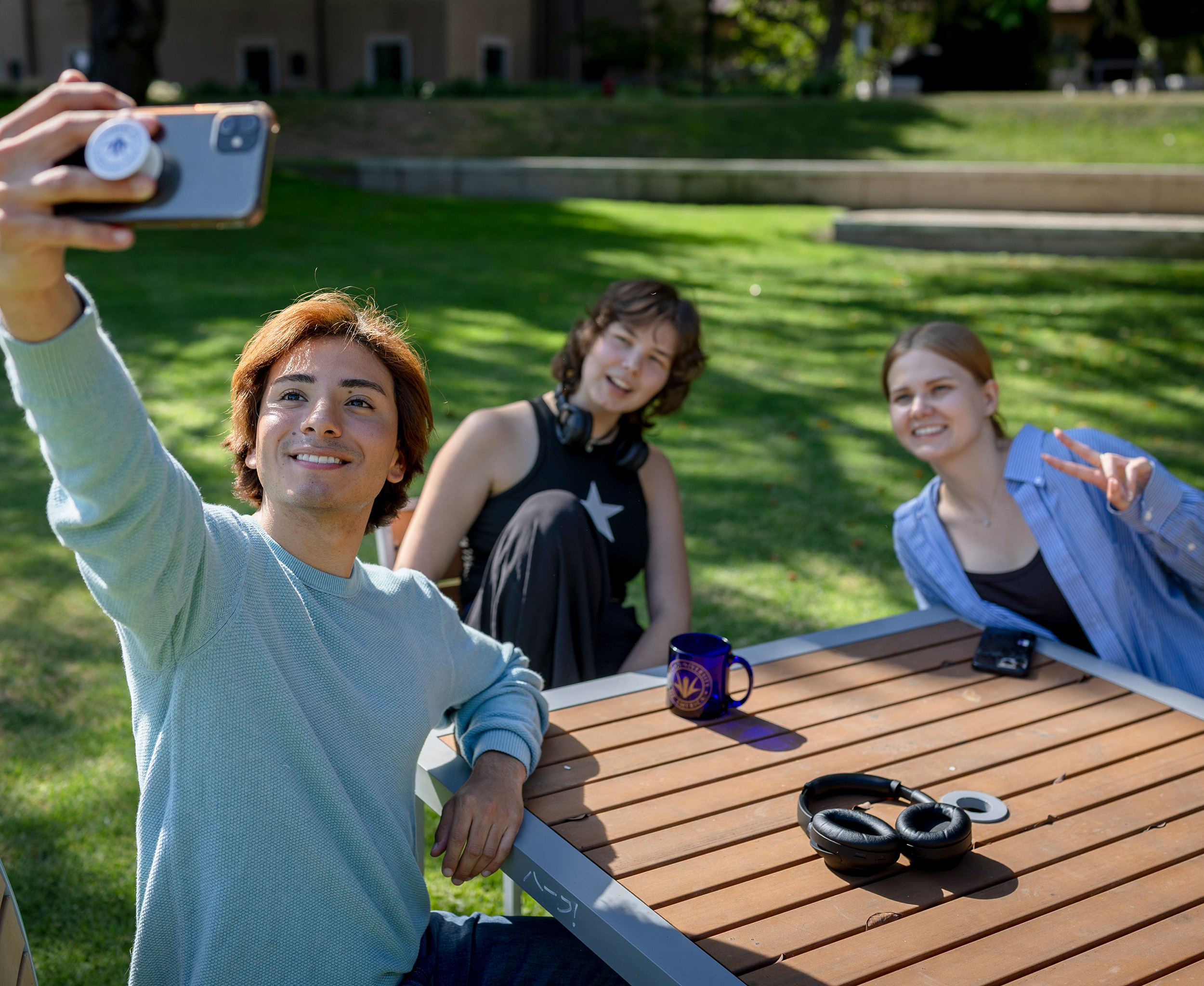  A young man takes a selfie with two friends while sitting at an outdoor wooden table on a sunny campus lawn.