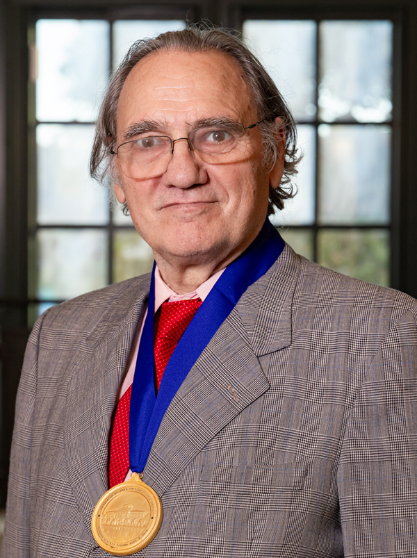 A grey-haired man with glasses poses for a headshot wearing a grey suit with a red tie and a gold medal with a blue band.