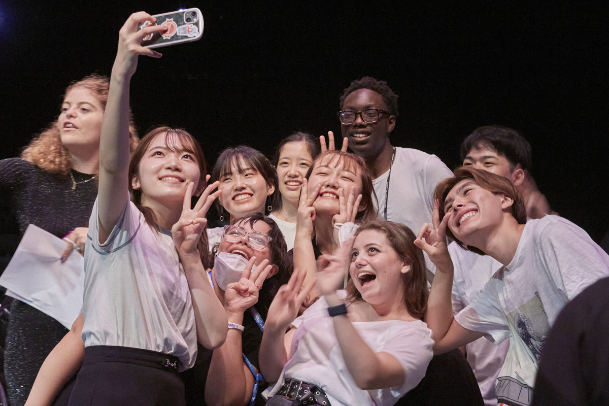 A group of students smile and hold up peace signs to the camera as they pose for a selfie.