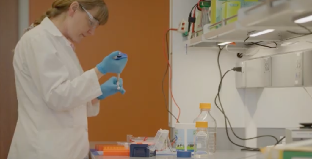 A professor in a white lab coat, safety glasses, and blue gloves uses a pipette to work with samples in a bright laboratory. In the background, lab equipment and glass bottles are visible on the workbench and shelves.