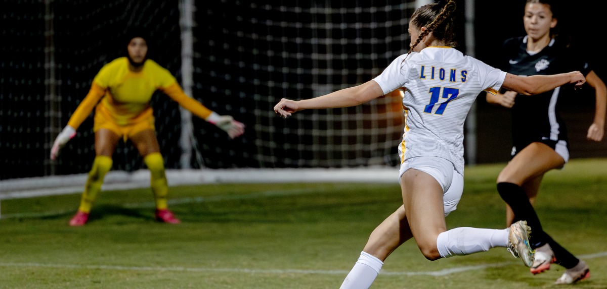 A women's soccer player is shooting on goal past a defender and goalkeeper.