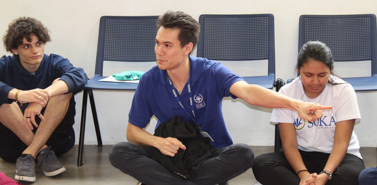 Scott Bower sits cross-legged on the floor in between two students and is pointing to his left