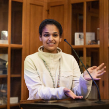 A woman wearing a white sweater and a lanyard speaks into a microphone and talks with her hands.