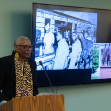 Gail E. Thomas wears a tiger-striped scarf and a black suit as she speaks at a lectern in front of a tv screen depicting the Greensboro sit-ins 