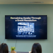 A man wearing a black turtleneck and grey dress pants speaks into a microphone while standing to the right of the tv as a woman wearing a light brown cardigan over a black shirt looks on