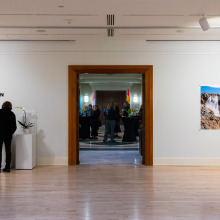 A woman and child sign the guestbook by the doorway of the gallery as attendees mingle in the atrium outside. Two large photographs are on a white wall to the right of the doorway.