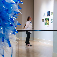 A woman wearing a white dress shirt and black pants stands with her arms behind her back as she views the gallery exhibit. A blue glass bottle art installation is in the foreground.