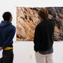 Two men stand in front of a large photograph of a mountainside. One of the men is standing with his arms behind his back and his hand is clutching papers.