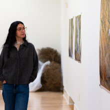 A brown-haired woman wearing a grey button up and blue jeans has her hands in her pockets as she walks alongside large photographs that are part of the exhibit.