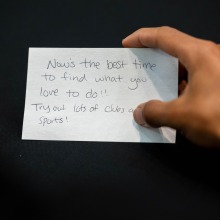 A hand holds a notecard reading "Now's the best time to find what you love to do!! Try out lots of clubs and sports!"