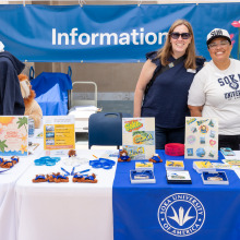 Two people stand behind an information desk that has Soka merchandise including sweatshirts, stickers, and ornaments.