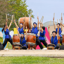 A group of ten people wearing royal blue tank tops and black pants stand with large wooden drums and raise their hands while holding drum sticks.