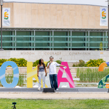 A male and a female student speak into microphones addressing the homecoming attendees while standing in front of large colorful and floral block letters that read "SOKA 25"