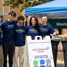 A group of four students and one staff member wearing matching navy t-shirts pose for a photo at their waste station, behind a sign instructing attendees.