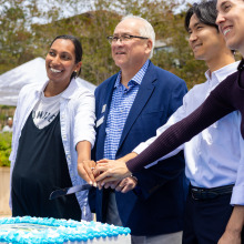 Four people put their hands together to jointly hold a knife for a cake-cutting photo op as they smile at another camera.