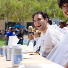 Multiple people wearing bibs smile and laugh as they sit at a table and prepare to participate in an eating competition.