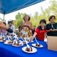 A group of students smile as they look at an select slices of chocolate cake from atop a table with a royal blue tablecloth.
