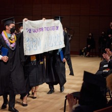 Graduates holding a sign saying, "We walk for the classes of 2020 and 2021" in honor of the graduating classes whose commencement ceremonies were cancelled due to Covid.