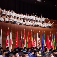 Members of the choir applaud for the graduates above a row of flags showing countries the graduates represent