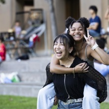 A girl wearing a black zipup shirt is giving another girl with spacebuns a piggyback ride as she holds up a peace sign. Both girls are smiling and laughing.