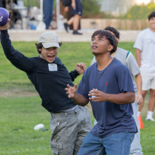 One student is dodging a ball while holding another dodgeball and another student in front of him is preparing to catch a ball