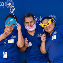 3 members of the facilities team wearing their blue polos hold up funny face masks during a shoot at the photobooth.