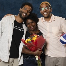 Lucas' mom and his two brothers pose in the digital media studio. His mother is holding a bouquet of flowers and one of his brothers is holding a volleyball.