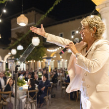 A female speaker with short curly hair, wearing a light cream-colored suit, speaks into a microphone