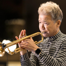A musician with gray hair wearing a black and white polka-dot shirt, playing a golden trumpet