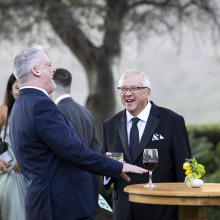 President E. Feasel talking with a man in suit while standing at a wooden cocktail table with drinksTwo men in suits laughing heartily while standing at a wooden cocktail table with drinks