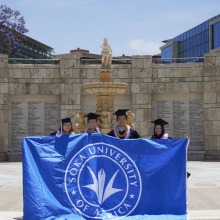 Four graduates pose with the Soka University of America banner in front of the Peace Lake Travertine Wall 