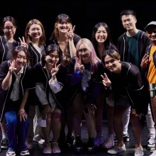 A group of ten students wearing jerseys smile and hold up peace signs on stage