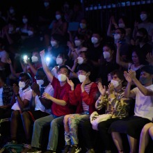 A large group of masked students are applauding and shining their cell phone lights.