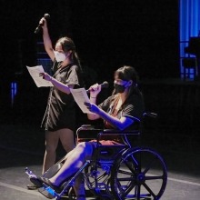 Two women wearing jerseys and masks are on stage reading from sheets of paper, into a microphone. The girl in the foreground is in a wheelchair and has a cast on her foot.