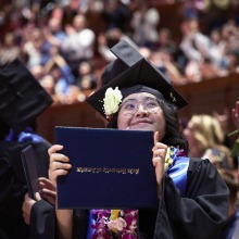 A proud graduate holds up her diploma and smiles.