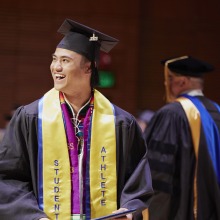A graduate wearing a "Student Athlete" sash smiles at the crowd during commencement