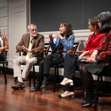 A panel of five speakers sit in a row of chairs. A brown haired woman in the middle is wearing a blue suit and gesturing with her hands as she speaks.