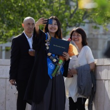 A graduate takes a selfie holding up her diploma with her family after commencement