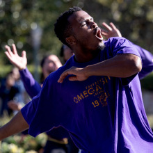 A close-up of a man in a purple "Omega Psi Phi" T-shirt performing with intense expression and outstretched arms during an outdoor event.