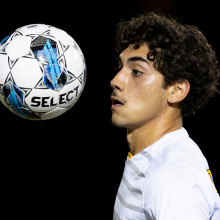 A close-up, high-action shot of a soccer player with curly hair frozen in time as a soccer ball floats inches from his face against a black background.