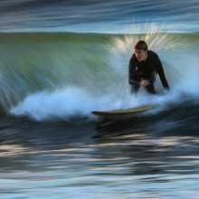 A surfer in a black wetsuit carves through a breaking wave, captured with a motion blur effect that creates streaks of white spray and blue water.