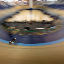An overhead long-exposure shot of a cyclist on a velodrome track, with the surrounding arena blurred into a circular zoom effect to emphasize speed.