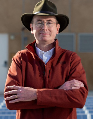 A man wearing a dark widebrimmed hat, glasses, and a red jacket with a white collar shirt underneath stands with his arms crossed and smiles at the camera.