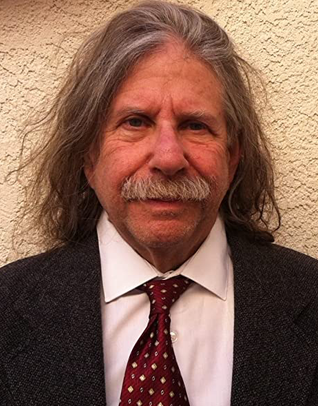 A man with gray long hair and a gray mustache is wearing a red polka dot tie and a black suit with a white shirt.