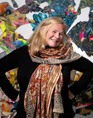 A blondehaired woman wearing a black longsleeve shirt and orange scarf stands with her hands on her hips in front of an abstract art display.