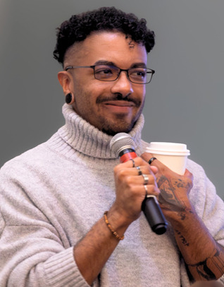 A man with short dark brown hair and glasses wearing a white turtleneck holds a microphone and coffee.