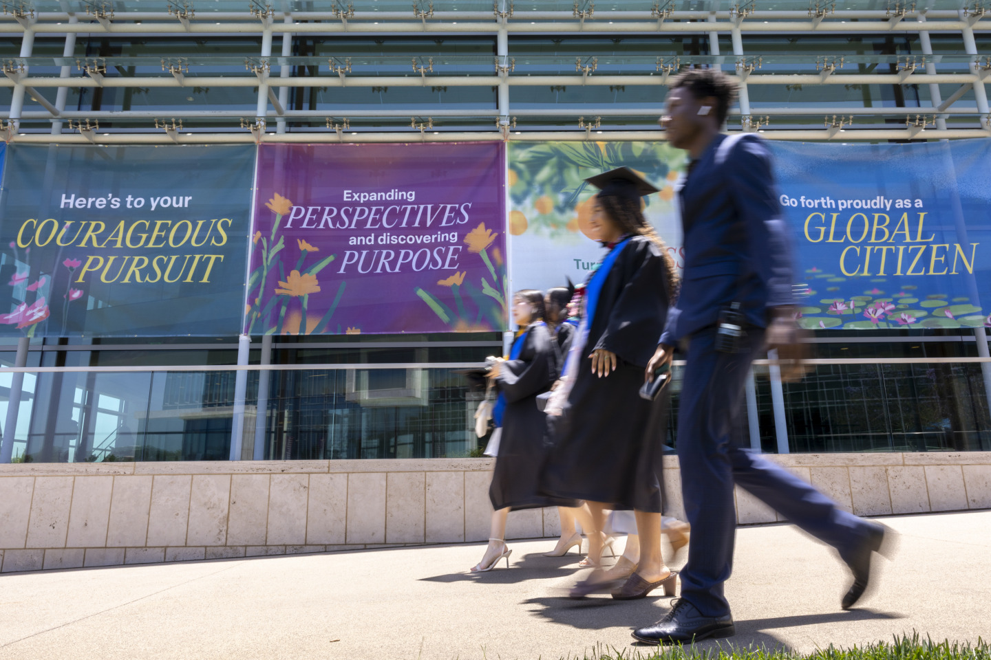Graduating students walk in a line wearing formal attire and their graduation gowns. They are walking alongside the Performing Arts Center which has branded window clings with mission-oriented language on them.